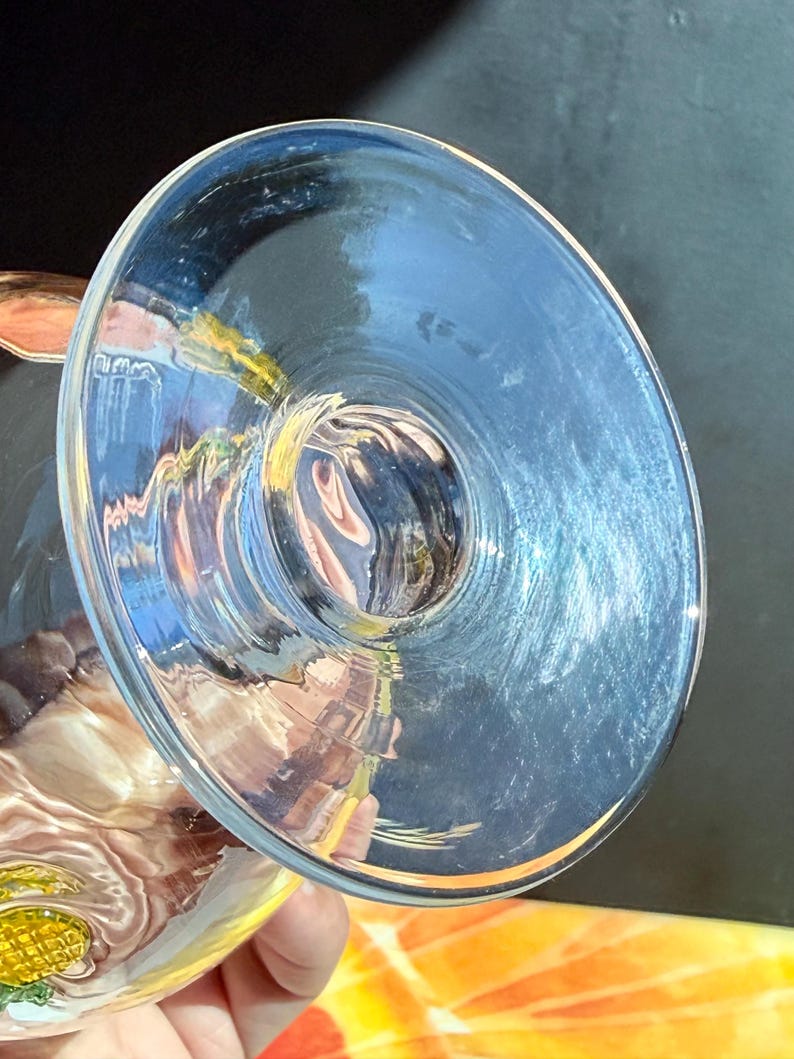 May include: A clear glass goblet with a wide, shallow bowl. The glass reflects light, creating a blue hue. The stem is not visible. The goblet is held by a person.