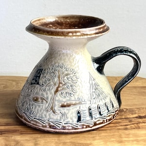 May include: A white ceramic pitcher with a brown rim and a blue handle. The pitcher has a raised design of a tree and a house.
