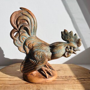 May include: A ceramic rooster figurine with a brown, green, and blue glaze. The rooster is standing on a wooden base.