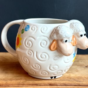 May include: White ceramic mug with a sheep design. The mug features a large handle and is decorated with embossed swirls and colorful accents, including a yellow sun and blue and yellow stars. Two sheep heads are on the side of the mug.