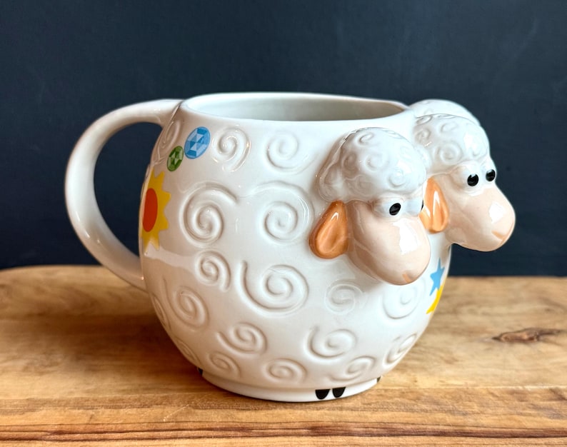 May include: White ceramic mug with a sheep design. The mug features a large handle and is decorated with embossed swirls and colorful accents, including a yellow sun and blue and yellow stars. Two sheep heads are on the side of the mug.