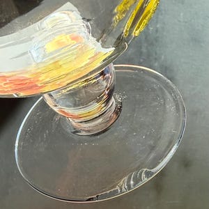 May include: A clear glass goblet with a wide, round base and a bulbous stem. The rim of the glass is decorated with yellow, orange, and green paint. The glass is partially filled with a clear liquid.