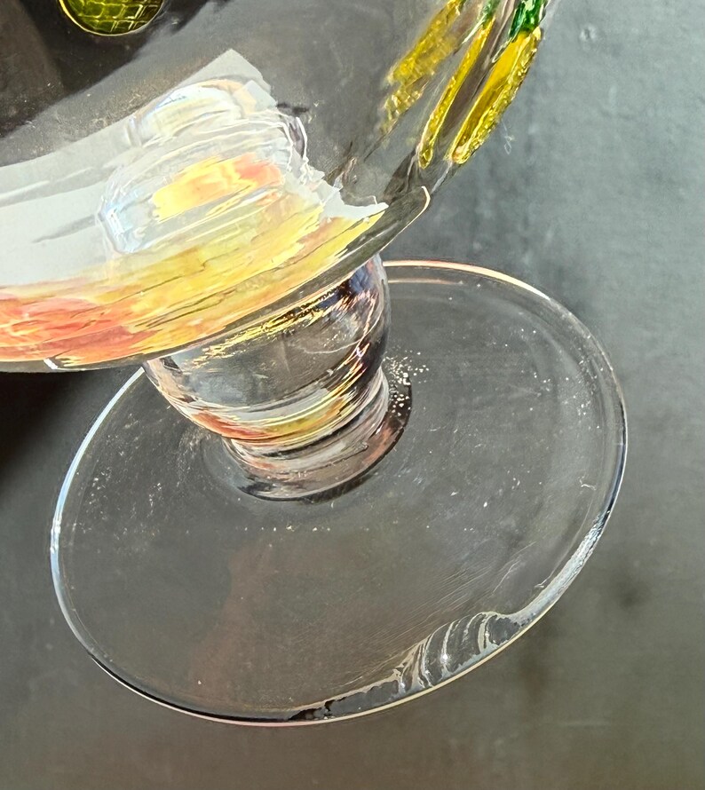 May include: A clear glass goblet with a wide, round base and a bulbous stem. The rim of the glass is decorated with yellow, orange, and green paint. The glass is partially filled with a clear liquid.