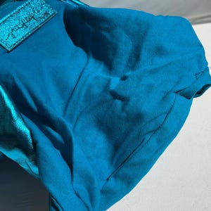May include: A turquoise handbag with a metallic blue accent. The bag has a zipper closure and a rectangular patch with cursive text. The fabric appears to be a sturdy cotton blend.