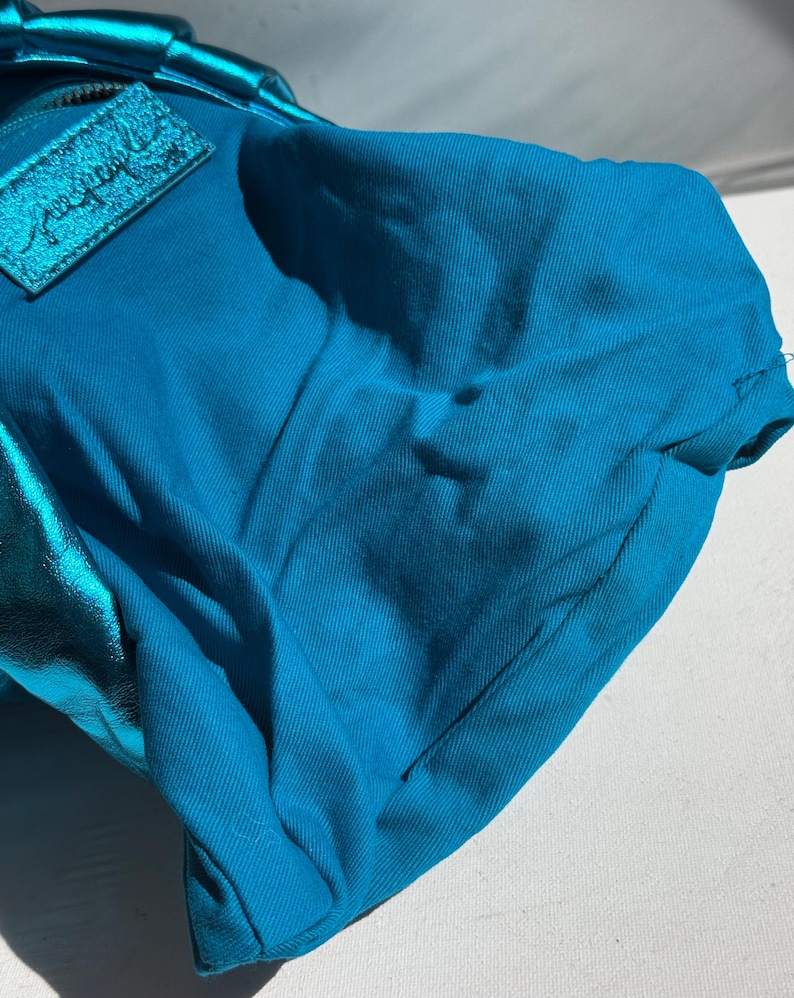 May include: A turquoise handbag with a metallic blue accent. The bag has a zipper closure and a rectangular patch with cursive text. The fabric appears to be a sturdy cotton blend.