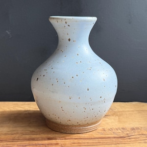 May include: A light blue ceramic vase with a speckled design. The vase has a rounded body, a narrow neck, and a flared rim. The base is a natural, unglazed color. The vase is sitting on a wooden surface.