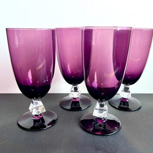 May include: Four elegant purple wine glasses with a dark base and a clear, geometric stem. The glasses have a tall, tulip-shaped bowl and are arranged on a dark surface.