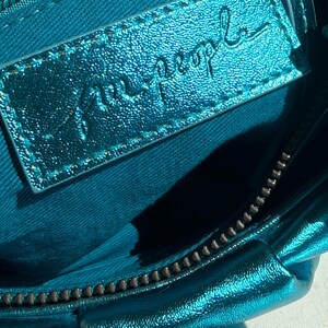 May include: Close-up of a teal-colored handbag interior. The bag features a metallic sheen and a zipper closure. A rectangular leather tag with the embossed text "free people" is visible.