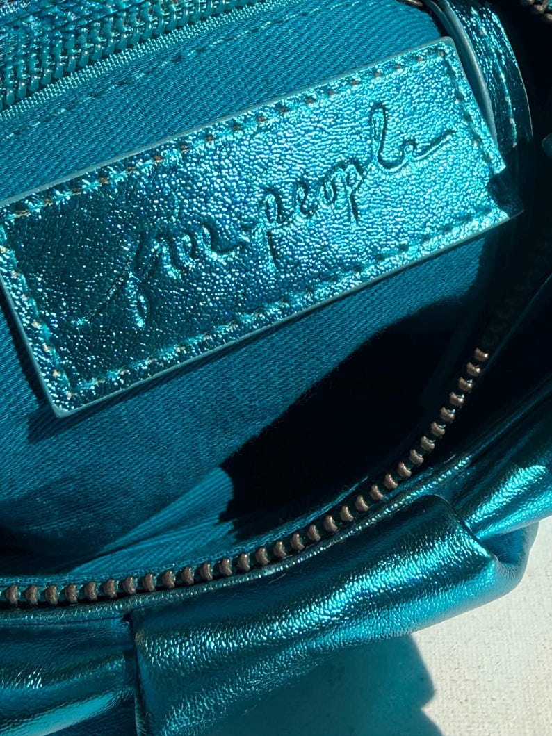 May include: Close-up of a teal-colored handbag interior. The bag features a metallic sheen and a zipper closure. A rectangular leather tag with the embossed text "free people" is visible.