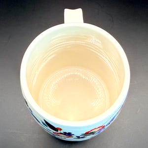 May include: Cream-colored ceramic mug with a floral design. The mug has a wide opening, a sturdy handle, and a light blue band with red, blue, and black floral patterns around the base. The interior is a light beige color.