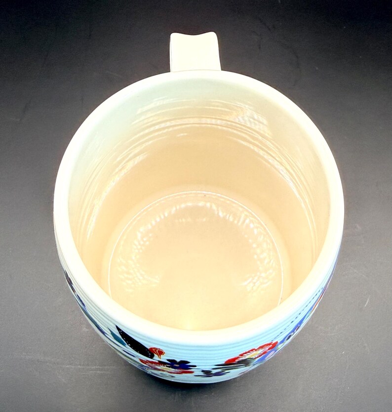 May include: Cream-colored ceramic mug with a floral design. The mug has a wide opening, a sturdy handle, and a light blue band with red, blue, and black floral patterns around the base. The interior is a light beige color.