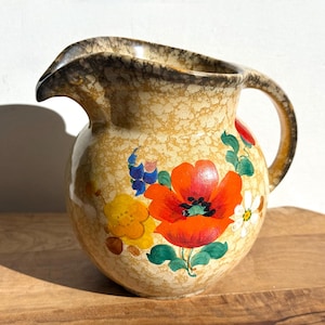 May include: A vintage ceramic pitcher with a tan speckled finish and a dark brown rim. The pitcher is adorned with hand-painted flowers in red, orange, yellow, blue, and white, with green leaves. It has a curved handle and a spout for pouring.