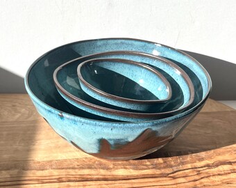 May include: Set of three nesting bowls in a teal blue glaze with a brown rim. The bowls are made of ceramic and have a rustic, handmade look.