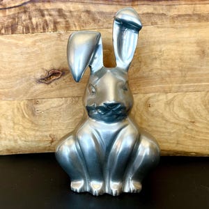May include: A silver-colored rabbit figurine with long ears and a detailed face. The bunny is sitting upright, with its front paws tucked in. The sculpture has a smooth, metallic finish and is set against a wooden background.
