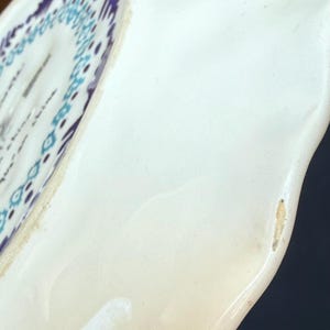 Puede incluir: Primer plano de un plato de cer&aacute;mica blanca con borde curvo. El plato presenta un patr&oacute;n geom&eacute;trico azul y morado en el borde. El plato tiene un acabado brillante y una forma ligeramente irregular.
