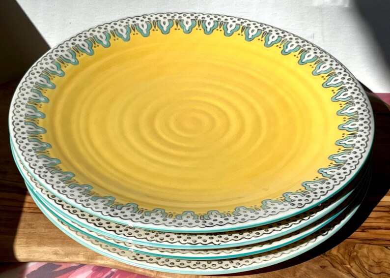 K&ouml;nnte beinhalten: Ein Set aus vier gelben Keramik-Dinnertellern mit einem blauen und wei&szlig;en Muster am Rand. Die Teller haben ein dezentes Spiraldesign in der Mitte.