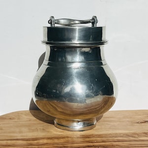 May include: A silver pewter jar with a lid and a handle. The jar has a rounded body and a wide mouth.