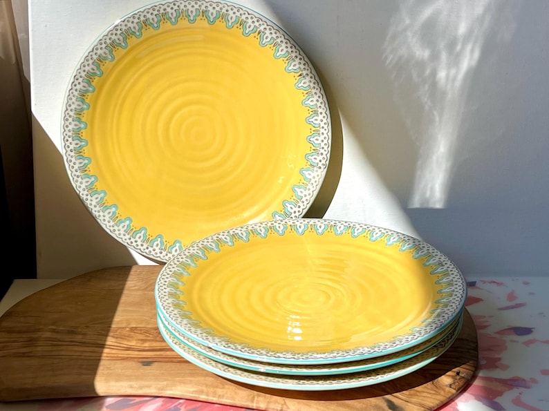 K&ouml;nnte beinhalten: Set aus vier gelben Keramik-Dinnertellern mit einem blauen und wei&szlig;en Muster am Rand. Die Teller haben eine geriffelte Textur.
