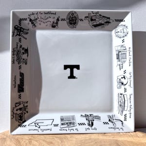 May include: A square white ceramic plate with a black "T" logo in the center. The border features black and white illustrations and text, including "Volunteers," "Neyland Stadium," and "Knoxville, Tennessee."