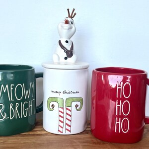 May include: Three ceramic mugs with holiday-themed designs. The green mug says "MEOWY & BRIGHT", the red mug says "HO HO HO", and the white mug says "merry christmas" with a green and red candy cane design. A snowman figurine sits on top of the white mug.