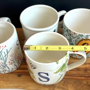 Pu&ograve; includere: Quattro tazze in ceramica bianca con manici sono disposte su una superficie di legno. Una tazza ha un motivo floreale con la lettera "S" e misura circa 10 cm di diametro. Altre tazze hanno vari disegni.