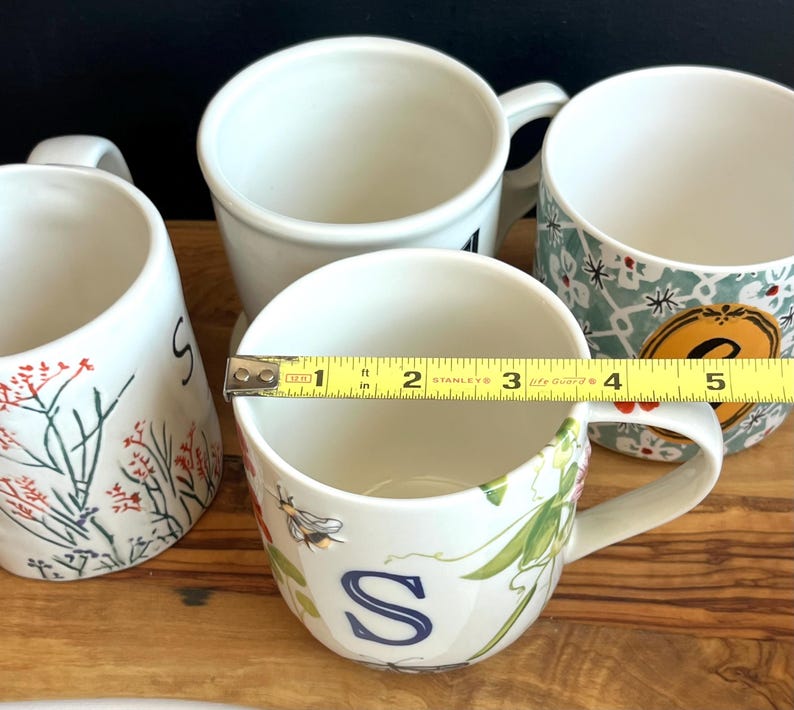 Pu&ograve; includere: Quattro tazze in ceramica bianca con manici sono disposte su una superficie di legno. Una tazza ha un motivo floreale con la lettera "S" e misura circa 10 cm di diametro. Altre tazze hanno vari disegni.