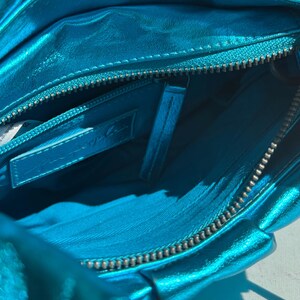 May include: Close-up of a turquoise metallic handbag, revealing the interior. The bag features a zipper closure and a small interior pocket. The lining is a matching turquoise color. The bag appears to be made of a soft, pliable material.