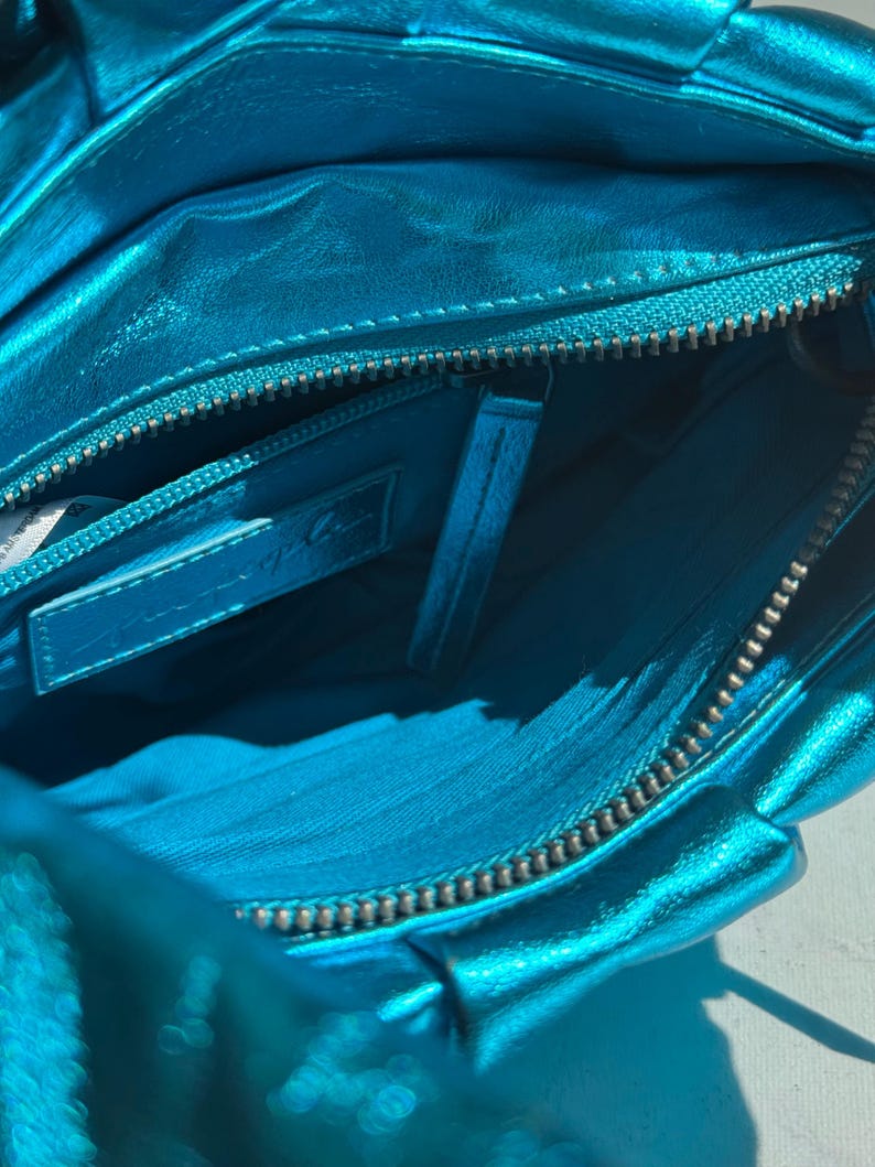May include: Close-up of a turquoise metallic handbag, revealing the interior. The bag features a zipper closure and a small interior pocket. The lining is a matching turquoise color. The bag appears to be made of a soft, pliable material.