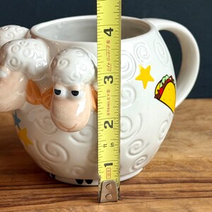 May include: White ceramic mug with a sheep design on the front and a handle on the side. The mug has a height of approximately 4 inches. It features a taco and star graphic on the side.