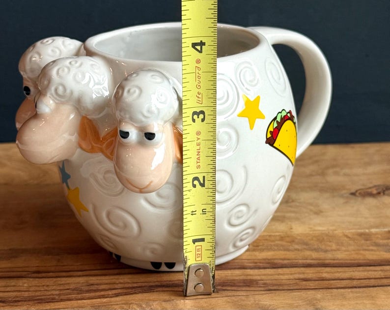May include: White ceramic mug with a sheep design on the front and a handle on the side. The mug has a height of approximately 4 inches. It features a taco and star graphic on the side.