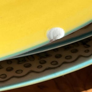 K&ouml;nnte beinhalten: Nahaufnahme einer gelben Keramikplatte mit einem kleinen wei&szlig;en Abplatzen am Rand. Der Teller ist auf einem anderen Teller mit einem blauen und wei&szlig;en Muster gestapelt.