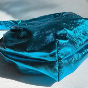 May include: A teal-colored handbag with a metallic sheen. The bag has a rectangular shape with a handle and is made of a shiny, textured material. The bag is sitting on a white surface.