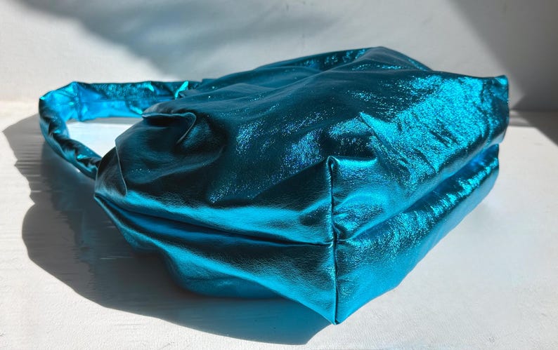 May include: A teal-colored handbag with a metallic sheen. The bag has a rectangular shape with a handle and is made of a shiny, textured material. The bag is sitting on a white surface.