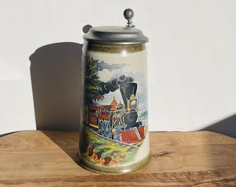 May include: A ceramic beer stein with a pewter lid features a vintage train engine pulling a string of boxcars. The train is depicted in a colorful scene with a forest in the background.