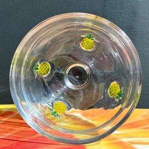 May include: Clear glass bowl with four yellow and green pineapple designs. The bowl has a circular shape with a dark center. The bowl is sitting on a colorful surface.