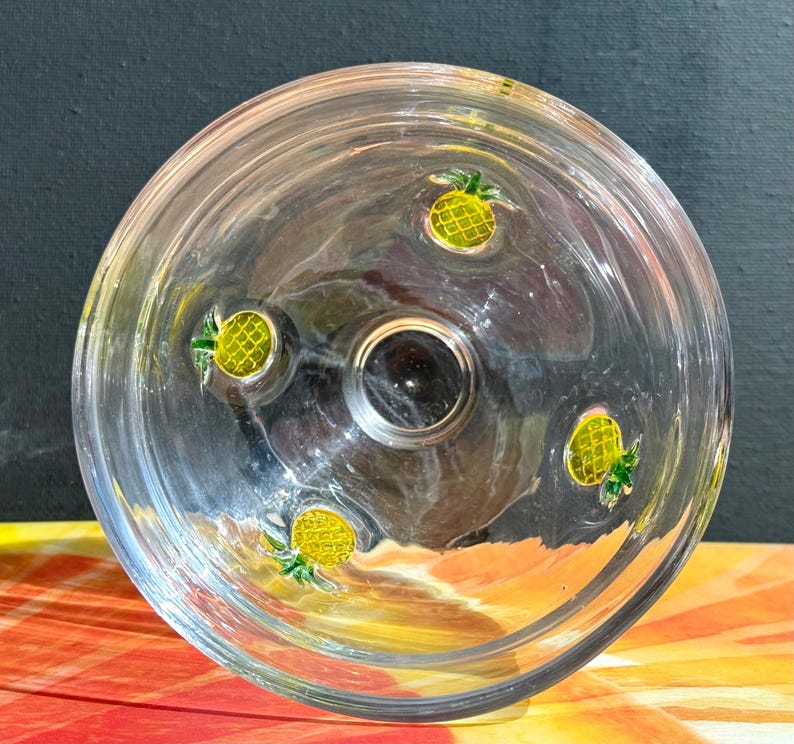 May include: Clear glass bowl with four yellow and green pineapple designs. The bowl has a circular shape with a dark center. The bowl is sitting on a colorful surface.