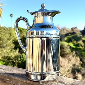 May include: A polished silver pitcher with a handle and a lid. The pitcher reflects the surrounding environment, including the sky and greenery. The design is classic and elegant, suitable for serving beverages.