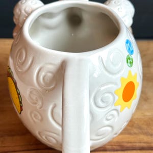 May include: White ceramic mug with a handle and a whimsical design. The mug features embossed swirls, a yellow sun, and a colorful taco illustration. The top of the mug has three decorative elements. The mug is on a wooden surface.