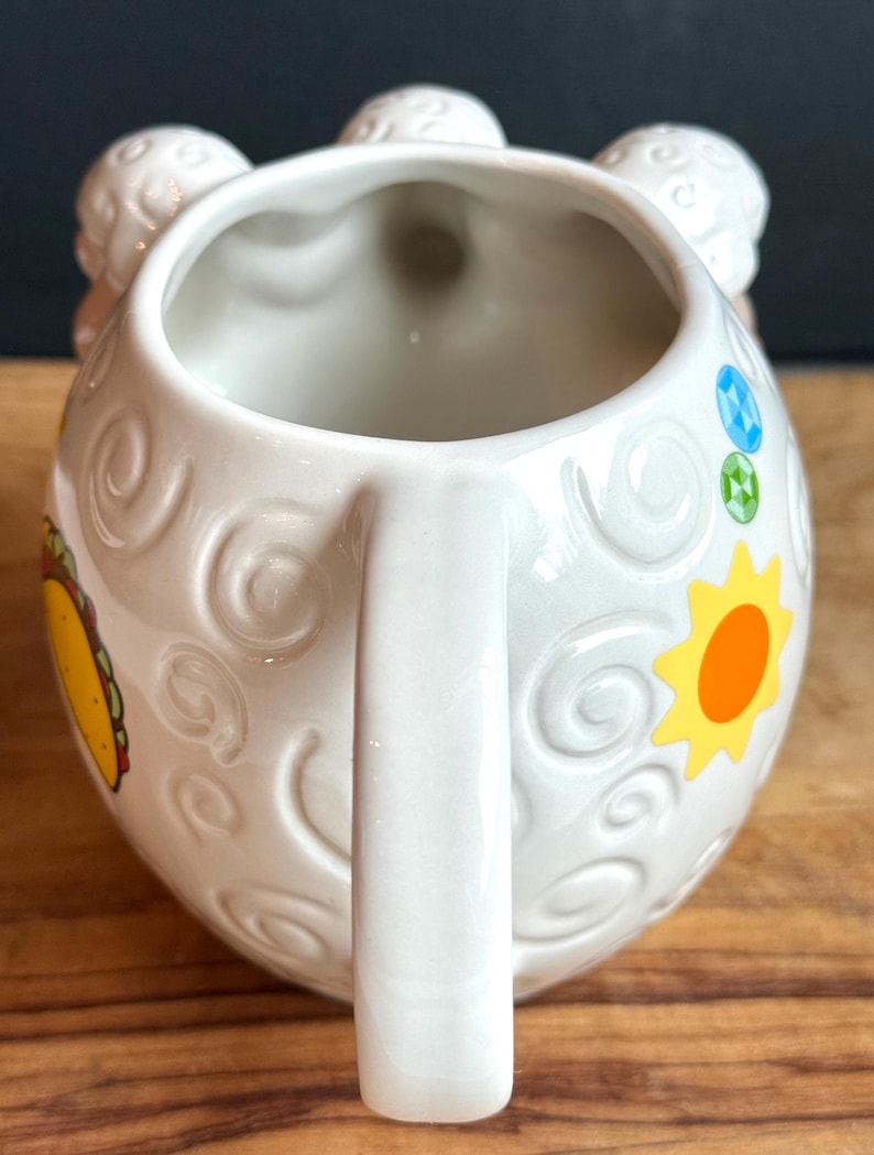 May include: White ceramic mug with a handle and a whimsical design. The mug features embossed swirls, a yellow sun, and a colorful taco illustration. The top of the mug has three decorative elements. The mug is on a wooden surface.