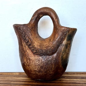 May include: A handcrafted, brown ceramic vase with a unique handle and a textured surface. The vase has a wide opening and a rounded base. The handle features a large, oval opening. The vase is displayed on a wooden tray.
