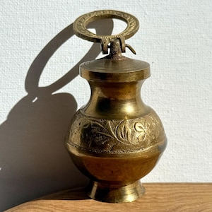 May include: A small, antique brass container with a decorative floral pattern. The container has a lid and a ring handle. The brass has a warm, golden hue. The container is likely used for storing small items or as a decorative piece.