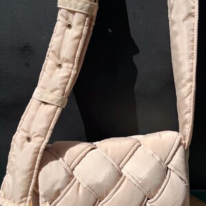 K&ouml;nnte beinhalten: Hellbeige Umh&auml;ngetasche mit gewebtem Design. Die Tasche hat einen breiten, verstellbaren Riemen mit goldfarbener Hardware. Die Tasche ist aus einem gesteppten Material gefertigt.