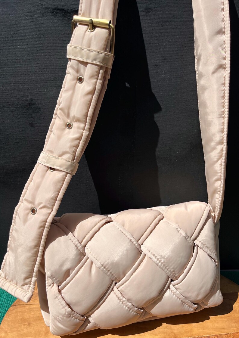 K&ouml;nnte beinhalten: Hellbeige Umh&auml;ngetasche mit gewebtem Design. Die Tasche hat einen breiten, verstellbaren Riemen mit goldfarbener Hardware. Die Tasche ist aus einem gesteppten Material gefertigt.