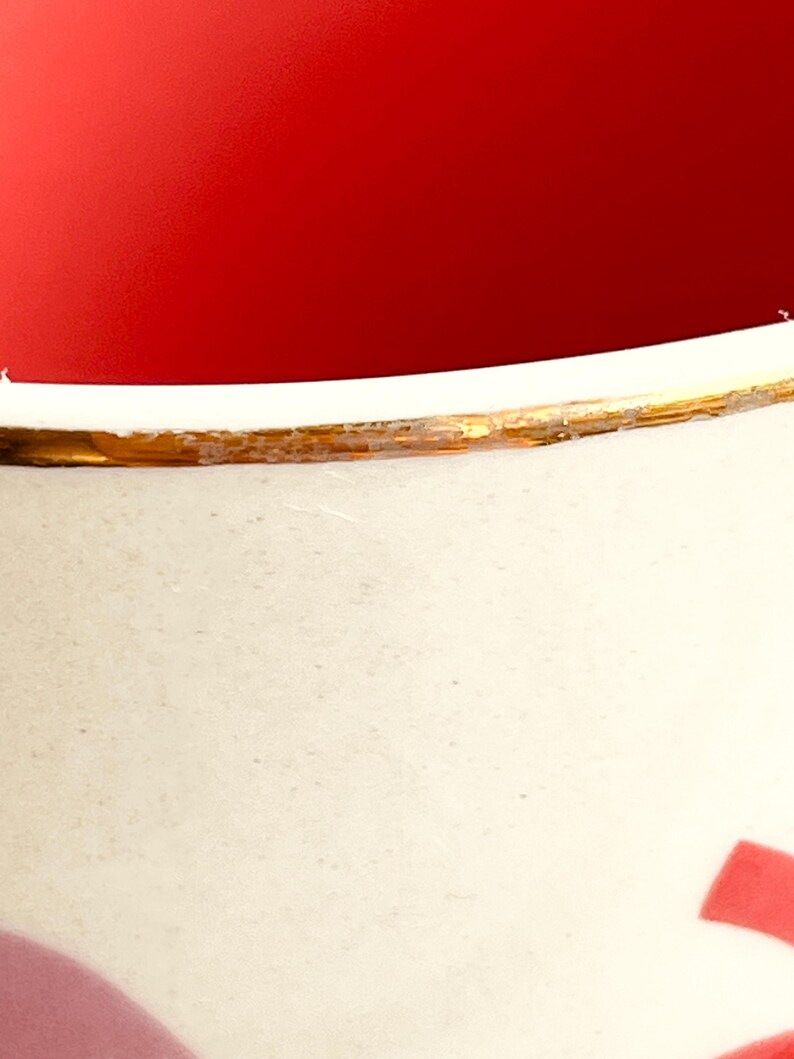 May include: A white ceramic mug with a gold rim and a red and pink design. The mug is empty and the inside is red.
