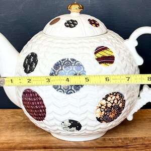 May include: White ceramic teapot with a textured surface and colorful, patterned circular designs. The teapot measures approximately 9 inches in length, including the handle and spout, and features a gold-colored finial on the lid.