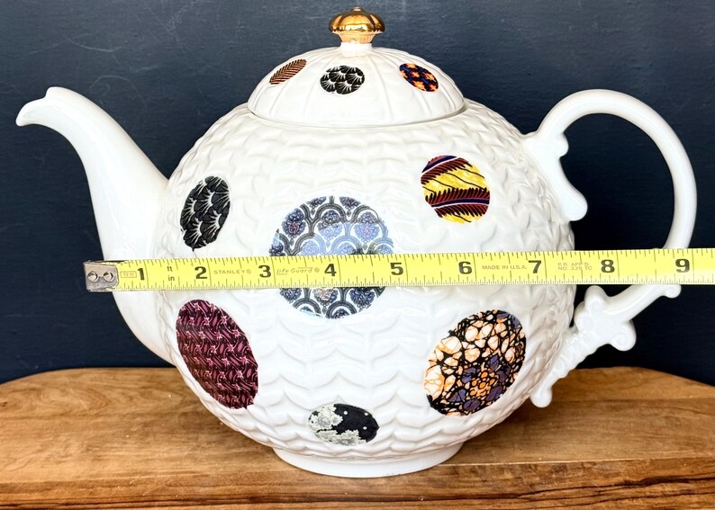 May include: White ceramic teapot with a textured surface and colorful, patterned circular designs. The teapot measures approximately 9 inches in length, including the handle and spout, and features a gold-colored finial on the lid.