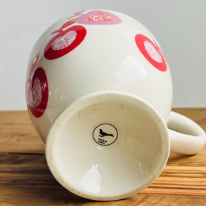 May include: White ceramic mug with a red and pink polka dot pattern. The mug has a handle and a black bird logo on the bottom that reads "Made in China."