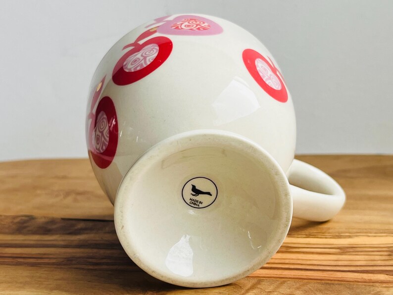 May include: White ceramic mug with a red and pink polka dot pattern. The mug has a handle and a black bird logo on the bottom that reads "Made in China."
