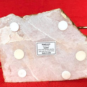 May include: A light pink, irregularly shaped stone slab with gold edging. The underside has six white circular pads and a label with text. The text reads "Weight Limit 6.008 kg" and "Made in India".