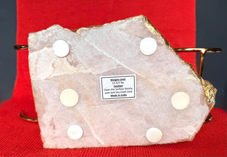 May include: A light pink, irregularly shaped stone slab with gold edging. The underside has six white circular pads and a label with text. The text reads "Weight Limit 6.008 kg" and "Made in India".